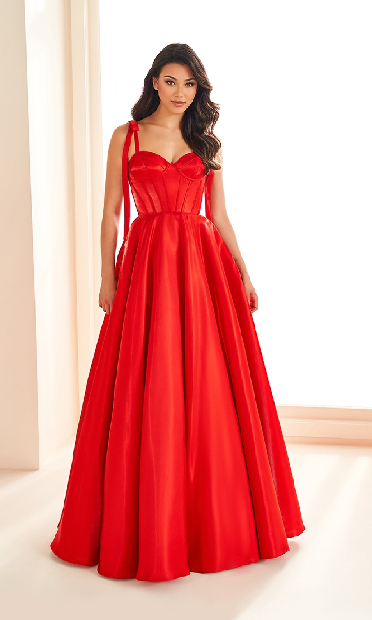 A woman stands indoors wearing the Ellie Wilde EW36210 Shoulder-Bows Prom Ball Gown, a floor-length, bright red sleeveless A-line gown with a fitted bodice and full skirt.